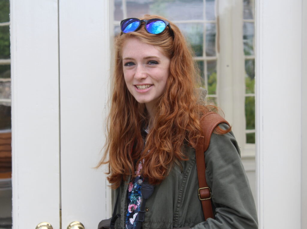 The writer stands with long, wavy, frizzy, red hair. It is messy and held back with sunglasses on the top of their head. 