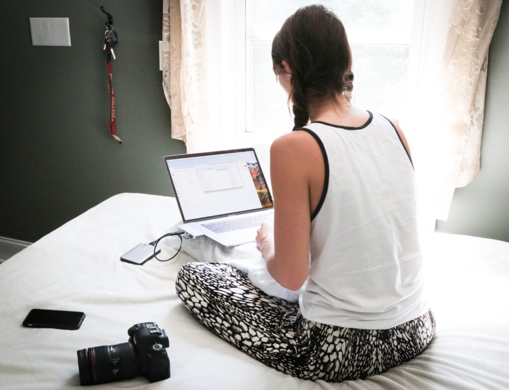 A young person with long braided hair sits on a bed with a laptop in their lap.