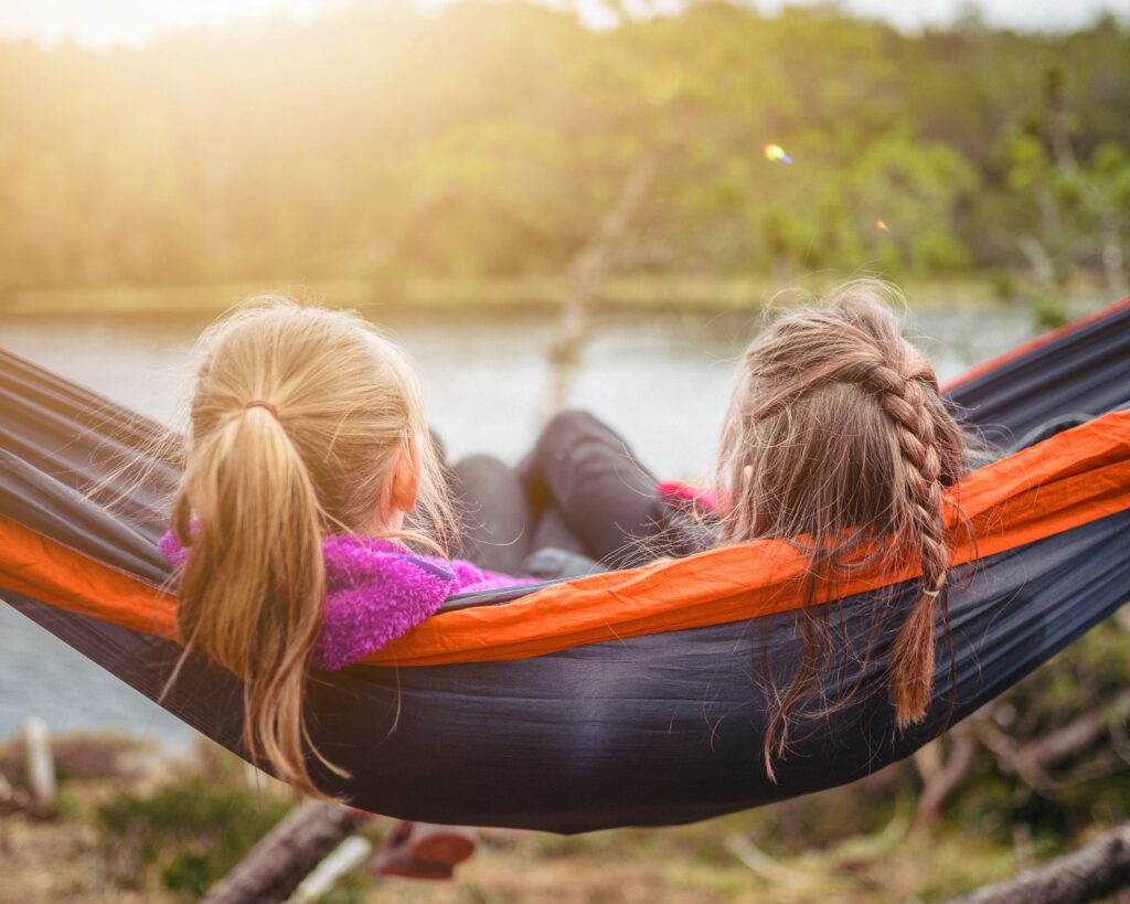 Two kids with long hair sit together in a hammock looking out at a lake.