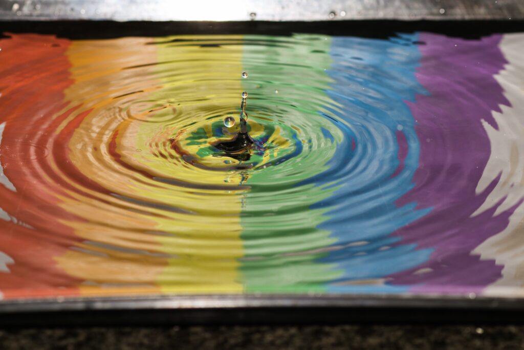 A pool of water reflects an lgbtq pride flag as a drop splashes in the middle, creating ripple effect.
