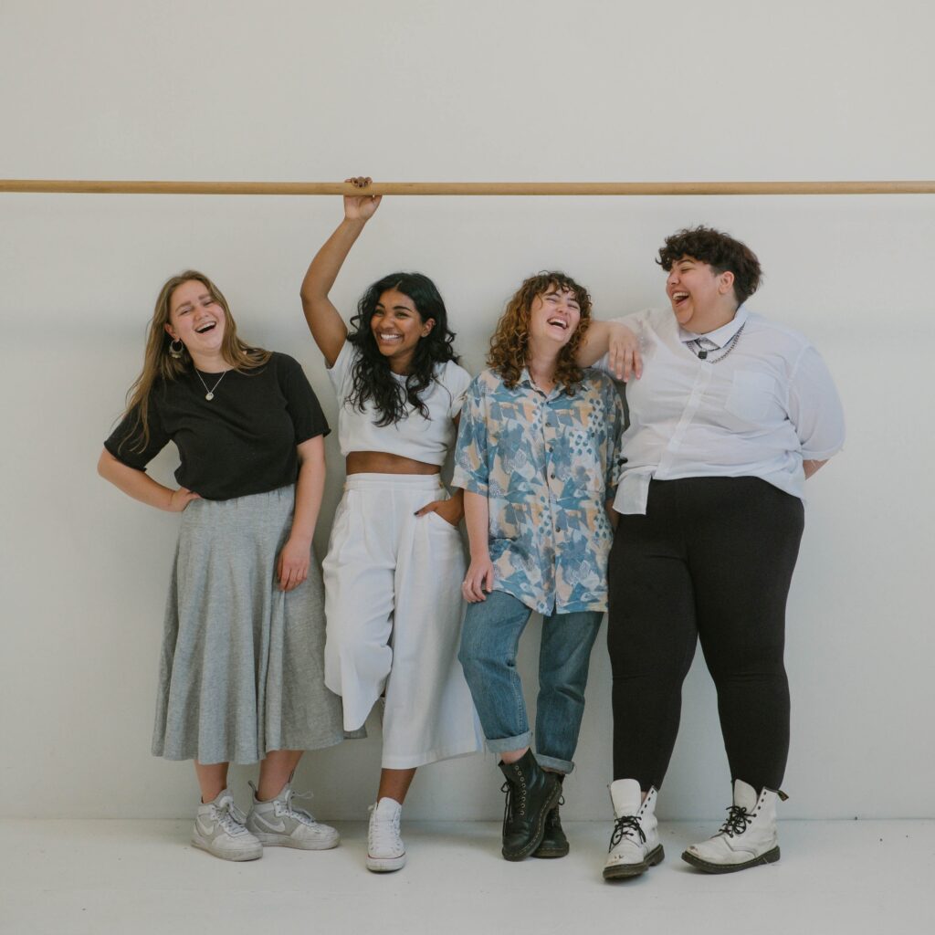Decorative. A group of 4 people have varying gender expressions and body types. From left to right the 4 people are as follows. 1. A person with long hair wears a plain tshirt with a long skirt and tennis shoes. 2. A thin person of color has long hair and wears a crop top with wide leg pants and converse. 3. A person with medium length curly hair wears a button down shirt with cuffed pants and black boots. 4. A fat person with a short haircut wears a button down shirt, necklaces, and black pants with white boots. 