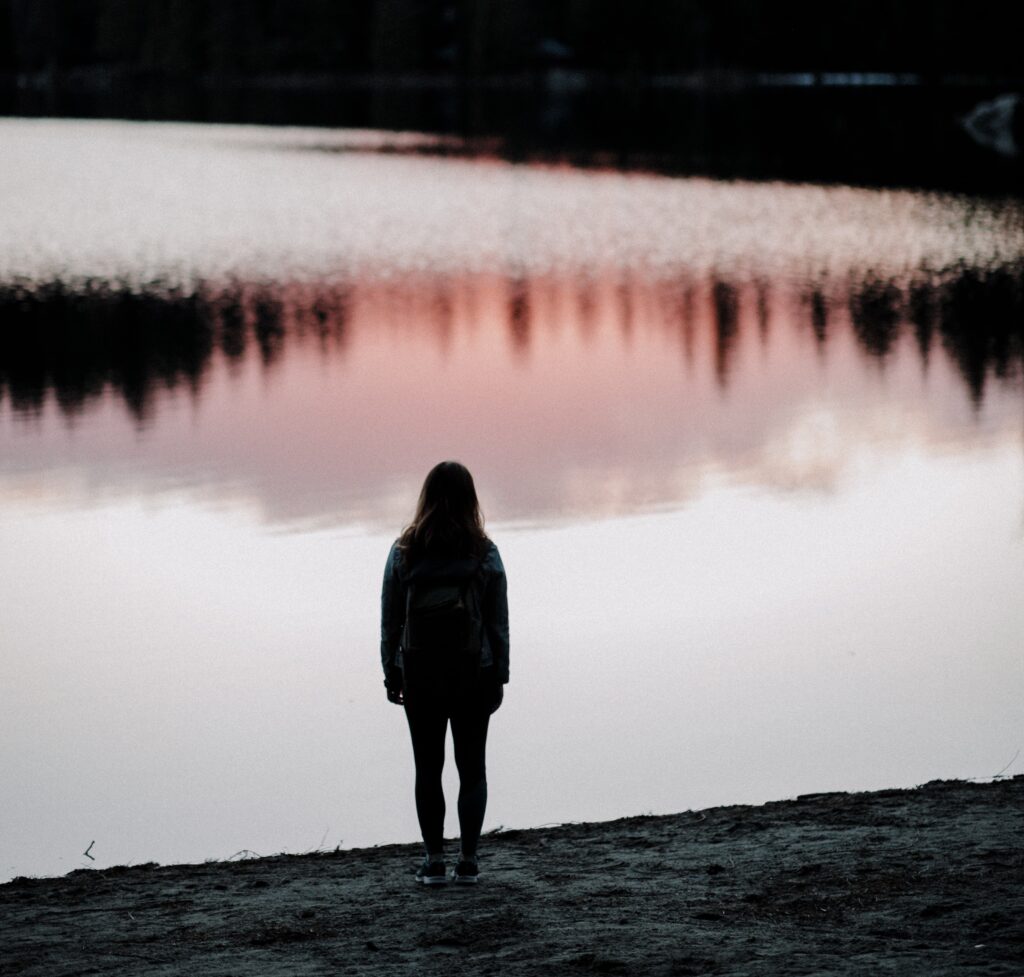 Decorative. A person with long hair stands near a still lake. The water's reflection reveals nearby forest and a pink sunset.